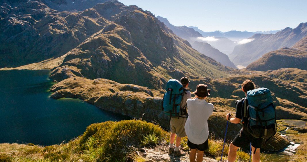 Three trampers looking out over a broad river valley and alpine lake from a mountain saddle